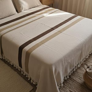 May include: Cream-coloured bedspread featuring brown and beige stripes, fringed edges, and matching pillows. The bed is made up with a neutral colour palette, creating a cosy and inviting atmosphere. The bedspread is made of a soft, textured fabric.