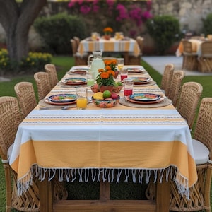 May include: An outdoor dining table set for a meal, covered with a yellow and white striped tablecloth. The table is set with colourful plates, glasses, and a bowl of fruit. Wicker chairs surround the wooden table, set on a lawn.