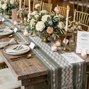 May include: A long wooden dining table features a sage green woven table runner with white geometric patterns and fringe ends. The table is set with floral centerpieces in brass bowls, tall taper candles in brass holders, votives, and menus.