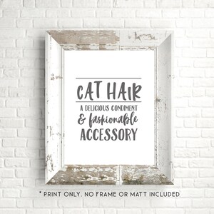 May include: A white framed print with the text "CAT HAIR A DELICIOUS CONDIMENT & fashionable ACCESSORY" in gray lettering. The print is on a white background and the frame is distressed white wood. The text "PRINT ONLY. NO FRAME OR MATT INCLUDED" is printed below the frame.