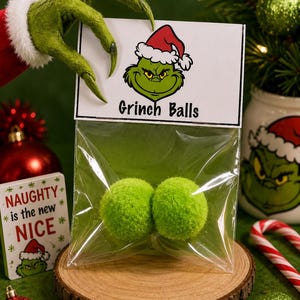 May include: A clear plastic bag containing two fuzzy lime green balls, topped with a white header card featuring a green character wearing a Santa hat and the text Grinch Balls in black font, presented as a festive holiday novelty item.