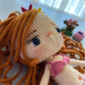 May include: A crocheted doll with long, brown hair, wearing a pink and white swimsuit. The doll has a sweet expression and is lying on a white surface.