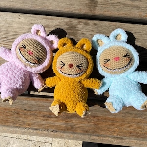May include: Three handmade crochet dolls in pastel colors: pink, yellow, and blue. Each doll has a smiling face with a stitched mouth and nose, and a hood with small ears. The dolls are lying on a wooden surface.