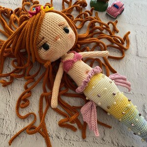 May include: A crocheted mermaid doll with long, curly orange hair, a pink bikini top, and a colourful tail with iridescent sequins. The doll is lying on a white brick surface.