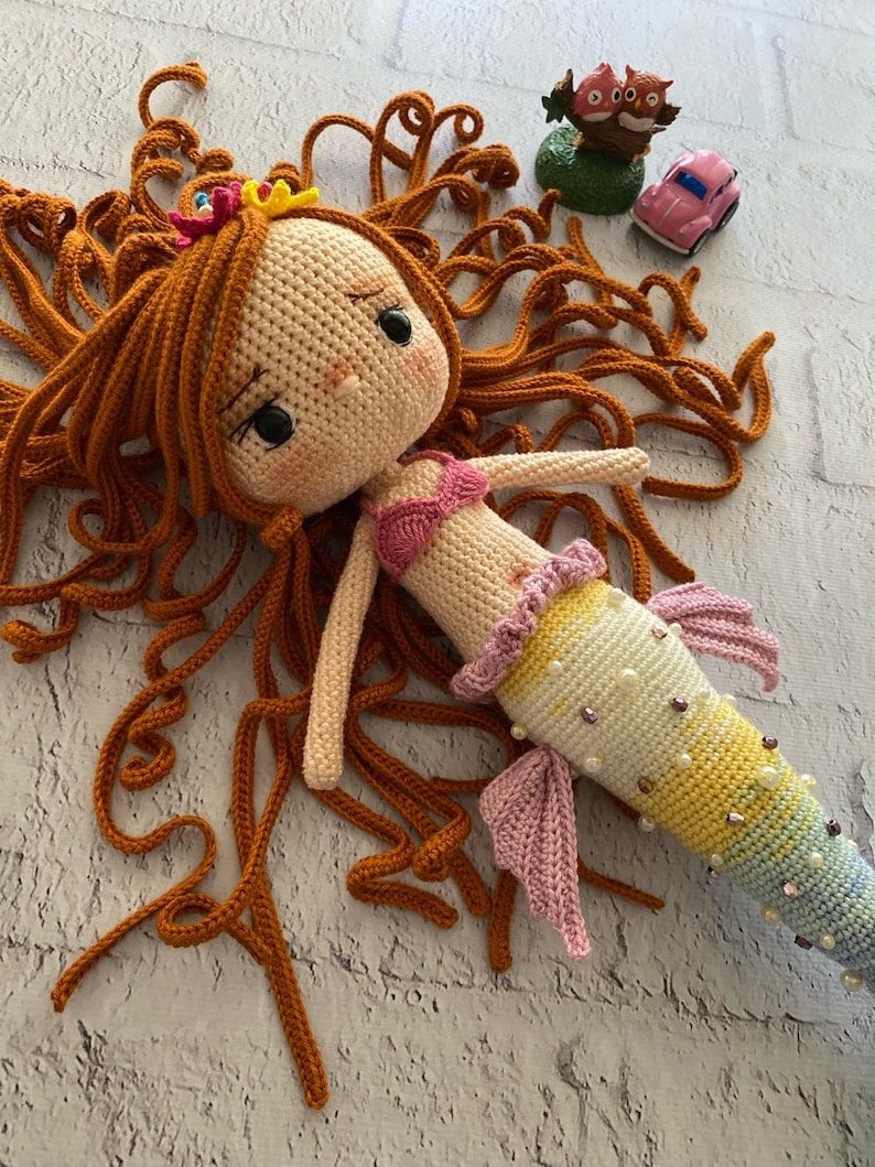 May include: A crocheted mermaid doll with long, curly orange hair, a pink bikini top, and a colourful tail with iridescent sequins. The doll is lying on a white brick surface.