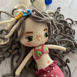 May include: A crocheted mermaid doll with gray hair, a pink and green swimsuit, and a pink tail. The doll is lying on a white surface.