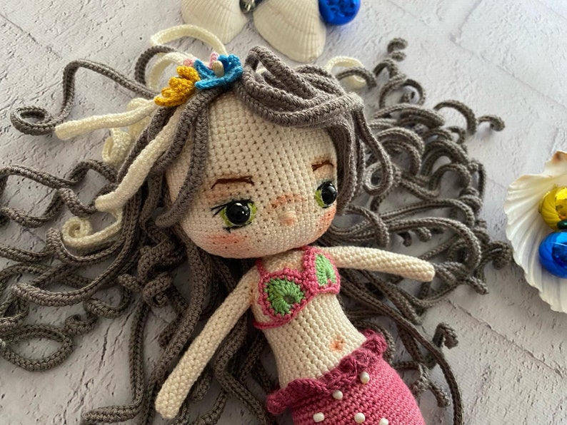 May include: A crocheted mermaid doll with gray hair, a pink and green swimsuit, and a pink tail. The doll is lying on a white surface.