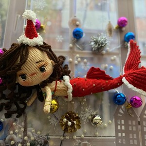 May include: A handmade crochet mermaid doll with brown hair, wearing a red and white Santa hat and a red tail. The doll is decorated with gold beads and white trim. Christmas ornaments in blue, pink, and silver surround the doll.