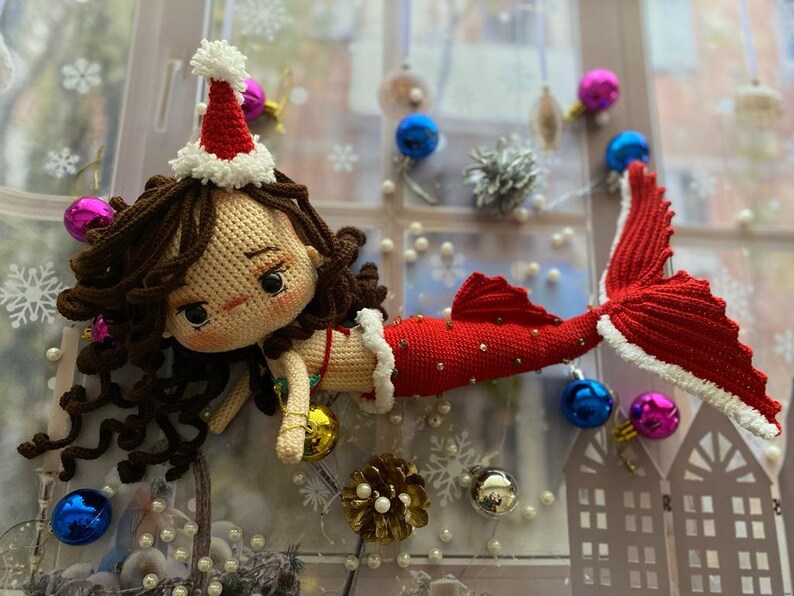May include: A handmade crochet mermaid doll with brown hair, wearing a red and white Santa hat and a red tail. The doll is decorated with gold beads and white trim. Christmas ornaments in blue, pink, and silver surround the doll.