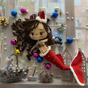 May include: A crocheted mermaid doll with brown hair, wearing a red and white Santa hat and a red tail. The doll is decorated with Christmas ornaments, including blue, pink, and gold baubles. The background includes a window with snowflake decorations.