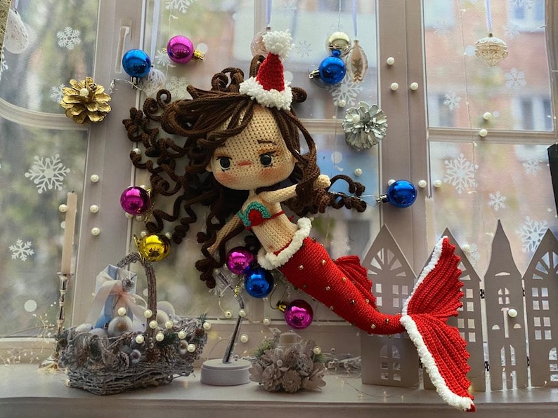 May include: A crocheted mermaid doll with brown hair, wearing a red and white Santa hat and a red tail. The doll is decorated with Christmas ornaments, including blue, pink, and gold baubles. The background includes a window with snowflake decorations.