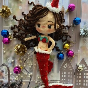 May include: A handmade crochet mermaid doll with brown hair, wearing a red tail and a Santa hat. The doll has a red and green top and is decorated with white trim. Christmas ornaments and decorations surround the doll.