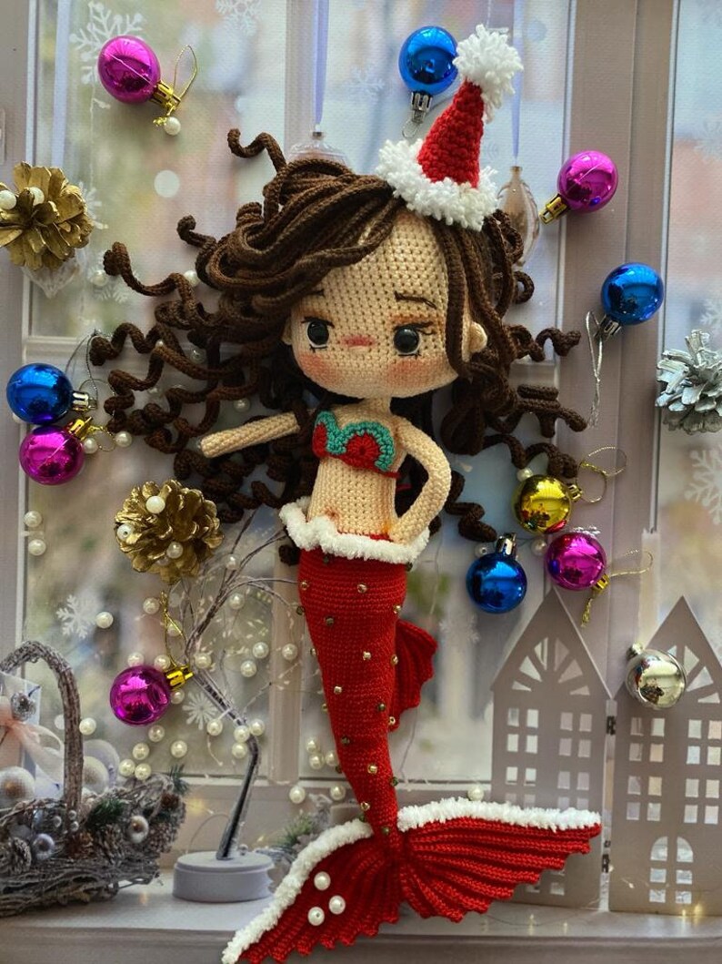 May include: A handmade crochet mermaid doll with brown hair, wearing a red tail and a Santa hat. The doll has a red and green top and is decorated with white trim. Christmas ornaments and decorations surround the doll.