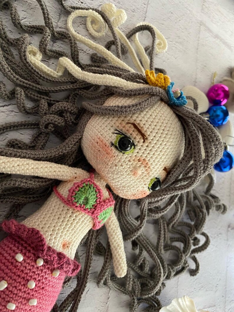 May include: A crocheted mermaid doll with long brown hair, a pink tail, and a green and pink bikini top. The doll has green eyes and a sweet expression.