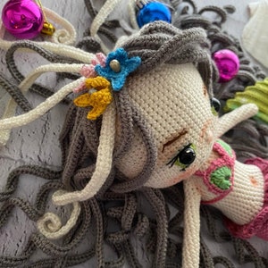 May include: A crocheted mermaid doll with long gray hair, a pink and green crocheted swimsuit, and a flower crown with blue, yellow, and white flowers.