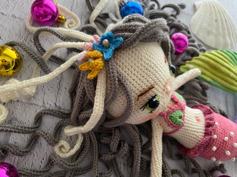 May include: A crocheted mermaid doll with long gray hair, a pink and green crocheted swimsuit, and a flower crown with blue, yellow, and white flowers.