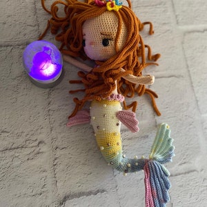 May include: A crocheted mermaid doll with long, curly, auburn hair, a yellow, pink, and blue tail, and a pearl-like embellishment. The doll is wearing a pink and yellow top and a flower crown.