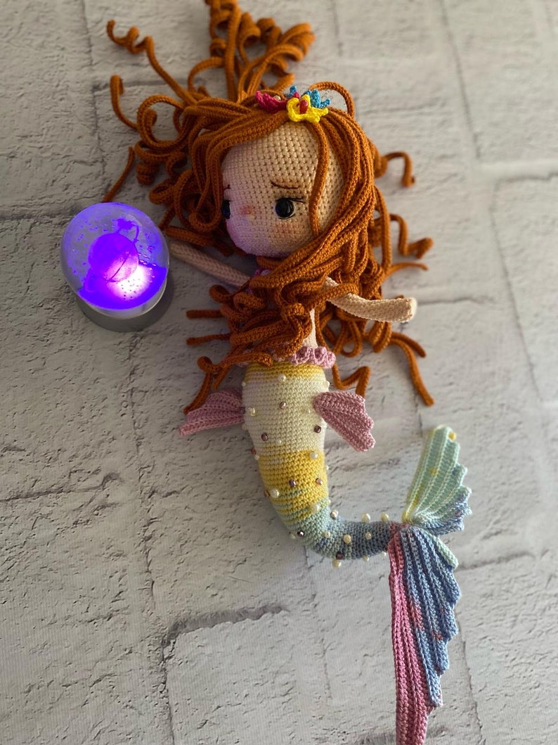 May include: A crocheted mermaid doll with long, curly, auburn hair, a yellow, pink, and blue tail, and a pearl-like embellishment. The doll is wearing a pink and yellow top and a flower crown.