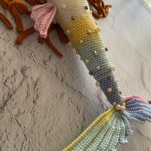 May include: A crocheted mermaid tail with a rainbow colour gradient. The tail is decorated with pearl and iridescent beads. The tail is made of yarn and is a handmade craft.