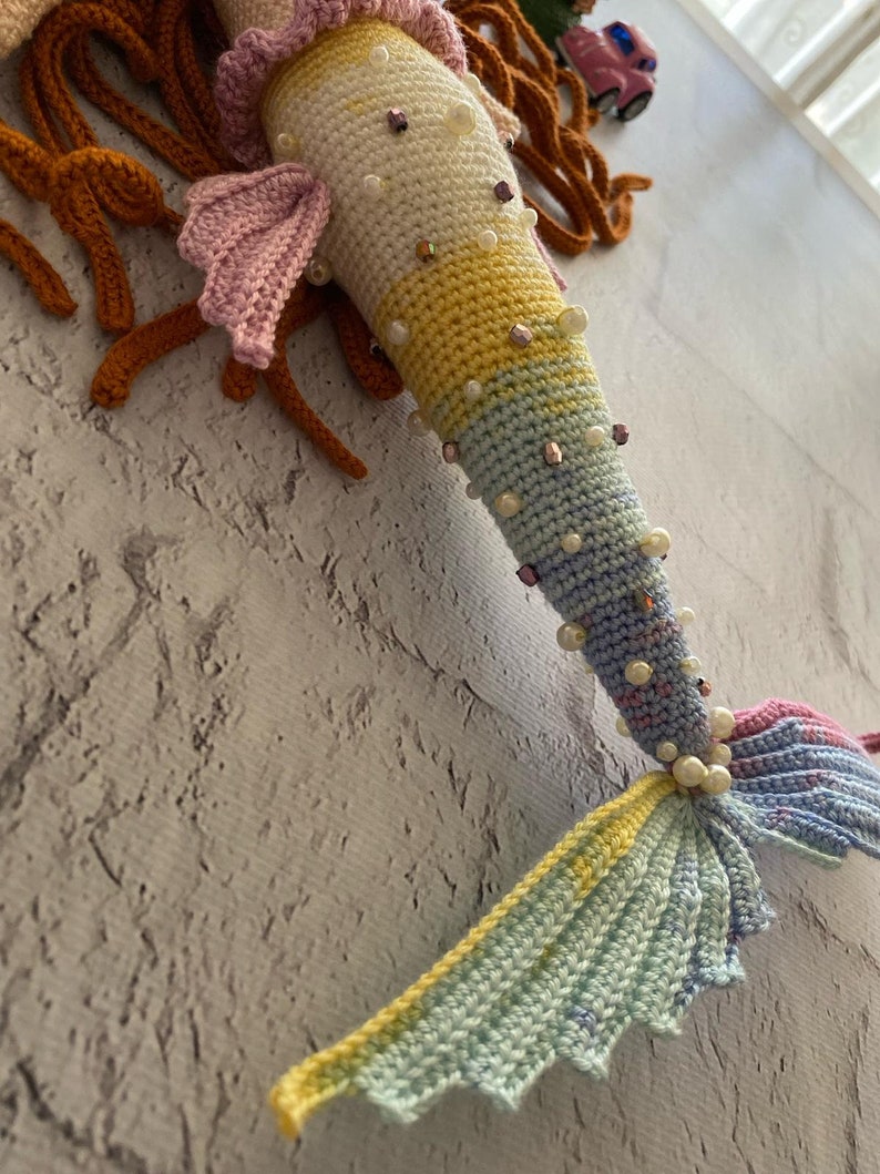 May include: A crocheted mermaid tail with a rainbow colour gradient. The tail is decorated with pearl and iridescent beads. The tail is made of yarn and is a handmade craft.