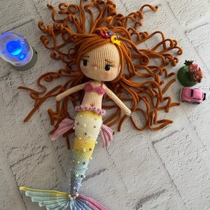 May include: A crocheted mermaid doll with long, curly orange hair, a pink top, and a colourful tail with pearl accents.
