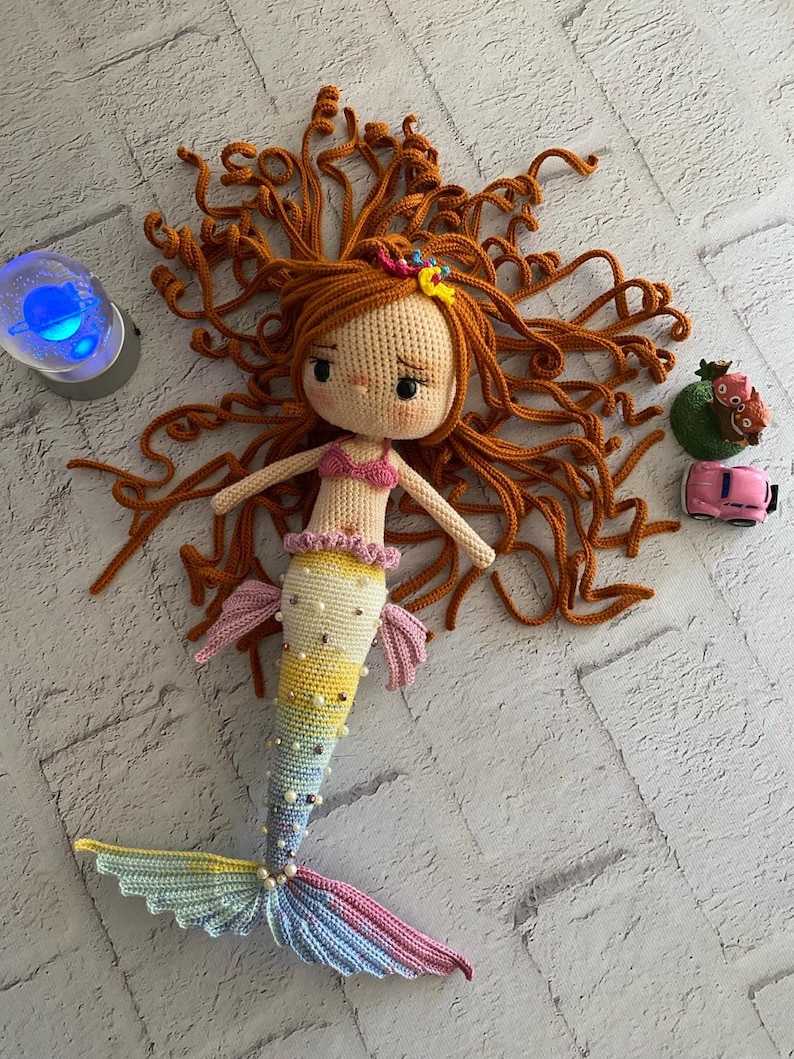 May include: A crocheted mermaid doll with long, curly orange hair, a pink top, and a colourful tail with pearl accents.