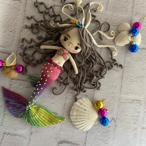 May include: A crocheted mermaid doll with long brown hair, a pink and purple tail, and a seashell crown. The doll is surrounded by seashells and colorful ornaments.