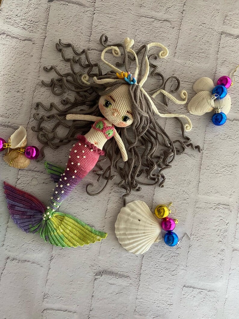 May include: A crocheted mermaid doll with long brown hair, a pink and purple tail, and a seashell crown. The doll is surrounded by seashells and colorful ornaments.