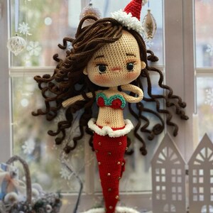 May include: A handmade crochet mermaid doll with brown curly hair, wearing a red and white Santa hat and a red tail with gold accents. The mermaid has a red and green top and is displayed near a window.