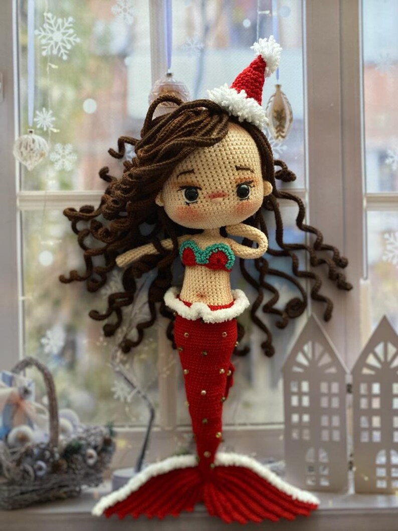May include: A handmade crochet mermaid doll with brown curly hair, wearing a red and white Santa hat and a red tail with gold accents. The mermaid has a red and green top and is displayed near a window.