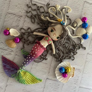 May include: A crocheted mermaid doll with long brown hair, a pink and white top, and a rainbow tail. The doll is lying on a white brick background with seashells and ornaments.