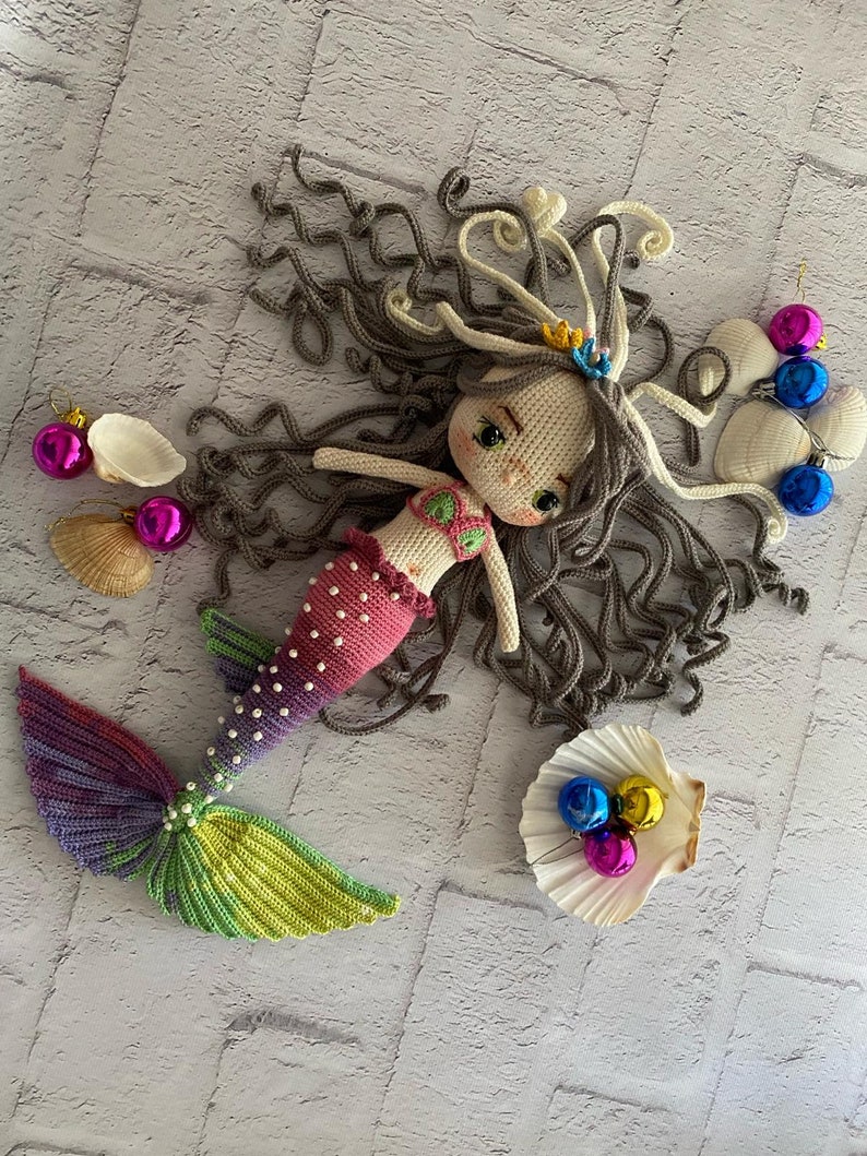 May include: A crocheted mermaid doll with long brown hair, a pink and white top, and a rainbow tail. The doll is lying on a white brick background with seashells and ornaments.