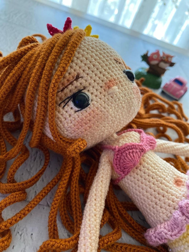 May include: A crocheted doll with long, brown hair, wearing a pink and white swimsuit. The doll has a sweet expression and is lying on a white surface.