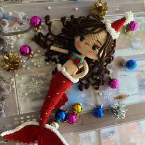 May include: A handmade crochet mermaid doll with brown hair and a Santa hat. The mermaid has a red tail with white trim and gold accents. The doll is surrounded by Christmas ornaments, including blue, pink, and gold baubles, and a gold pinecone.