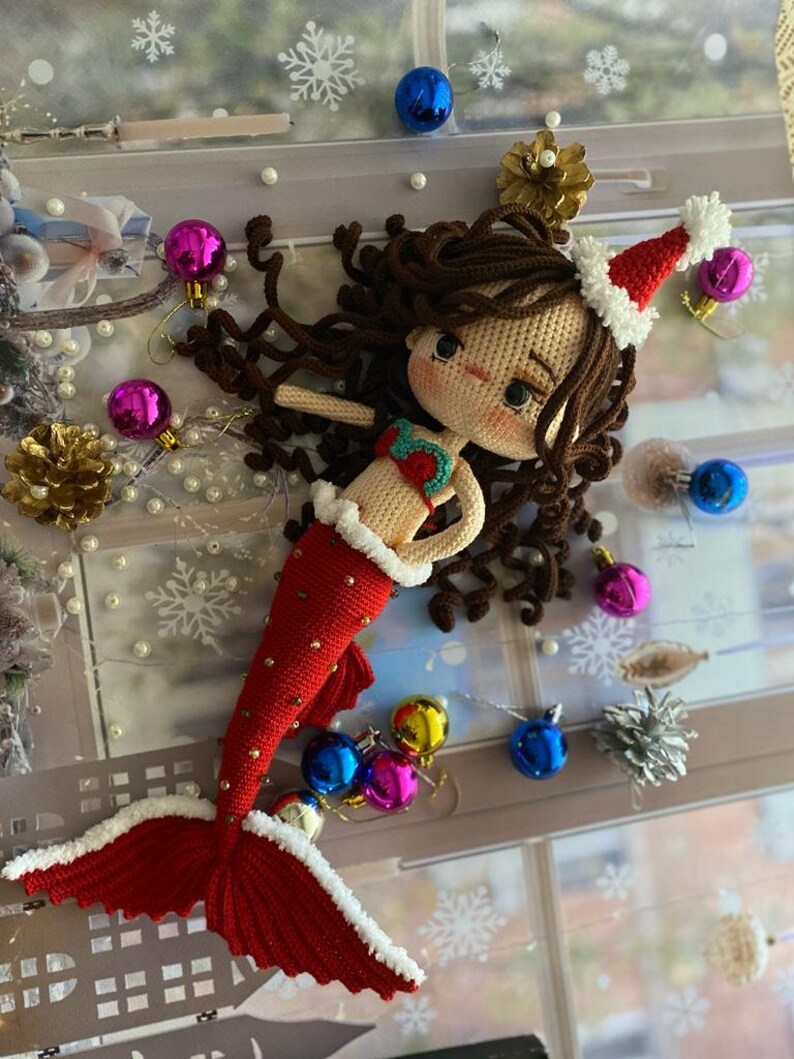 May include: A handmade crochet mermaid doll with brown hair and a Santa hat. The mermaid has a red tail with white trim and gold accents. The doll is surrounded by Christmas ornaments, including blue, pink, and gold baubles, and a gold pinecone.