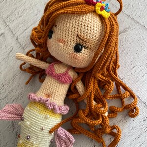 May include: A crocheted mermaid doll with long, curly orange hair, a pink bikini top, and a colourful tail with pearl accents.