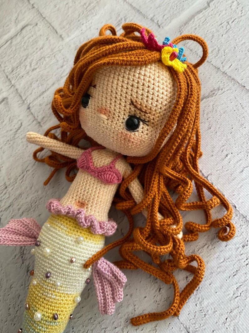 May include: A crocheted mermaid doll with long, curly orange hair, a pink bikini top, and a colourful tail with pearl accents.