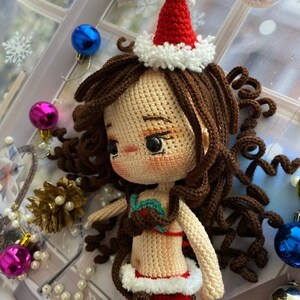 May include: A crocheted mermaid doll with brown curly hair, wearing a red and white Santa hat and a red mermaid tail. The doll has a tan complexion and is adorned with a red and green top. Christmas ornaments and decorations surround the doll.