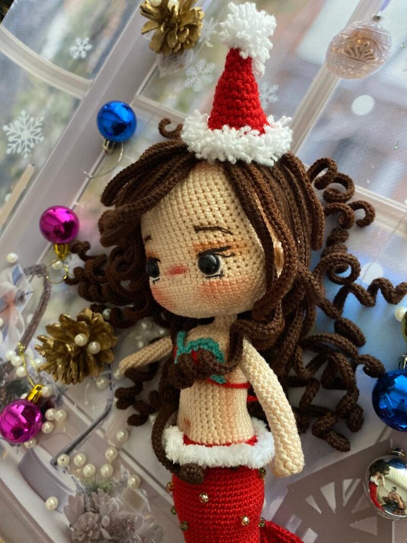 May include: A crocheted mermaid doll with brown curly hair, wearing a red and white Santa hat and a red mermaid tail. The doll has a tan complexion and is adorned with a red and green top. Christmas ornaments and decorations surround the doll.
