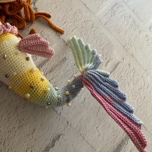 May include: A crocheted mermaid tail with a pastel rainbow colour scheme. The tail is decorated with pearl beads and iridescent sequins.