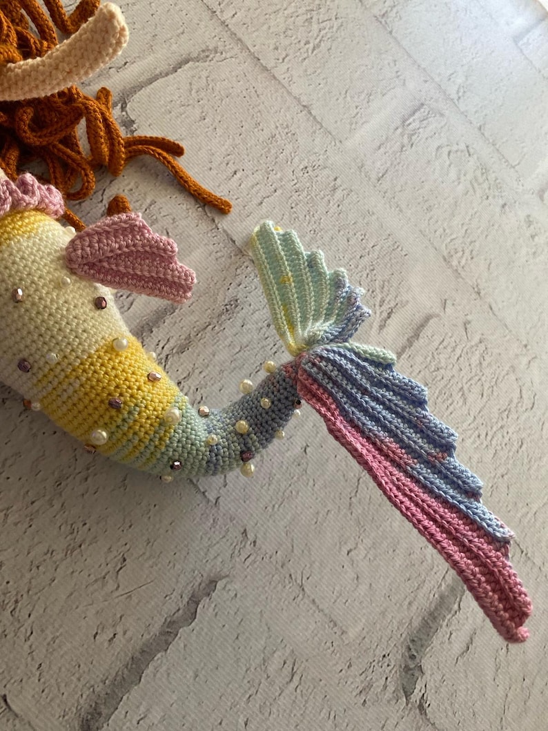 May include: A crocheted mermaid tail with a pastel rainbow colour scheme. The tail is decorated with pearl beads and iridescent sequins.