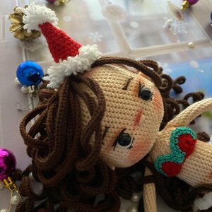 May include: A handmade crochet doll with brown hair and a Santa hat. The doll is wearing a red and white outfit with a green and red accent. Christmas ornaments and decorations surround the doll.