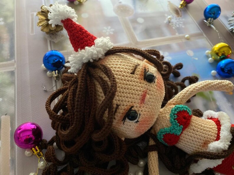 May include: A handmade crochet doll with brown hair and a Santa hat. The doll is wearing a red and white outfit with a green and red accent. Christmas ornaments and decorations surround the doll.