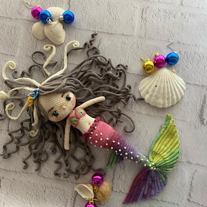 May include: A crocheted mermaid doll with long brown hair, a pink and white tail, and a green and purple fin. The doll is lying on a white brick background with seashells and colorful ornaments.