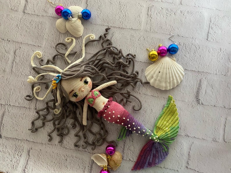 May include: A crocheted mermaid doll with long brown hair, a pink and white tail, and a green and purple fin. The doll is lying on a white brick background with seashells and colorful ornaments.