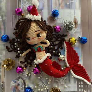 May include: A crocheted mermaid doll with brown hair, wearing a red and white Santa hat and a red tail. The mermaid is decorated with Christmas ornaments in a window setting. The mermaid's top is green and red.