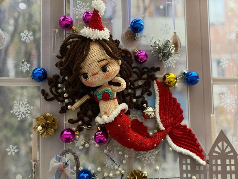 May include: A crocheted mermaid doll with brown hair, wearing a red and white Santa hat and a red tail. The mermaid is decorated with Christmas ornaments in a window setting. The mermaid's top is green and red.