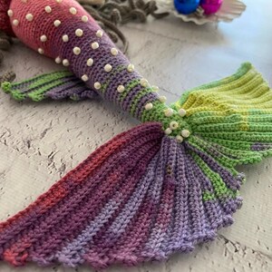 May include: A crocheted mermaid tail with a pink, purple, and green color scheme. The tail is decorated with white beads.