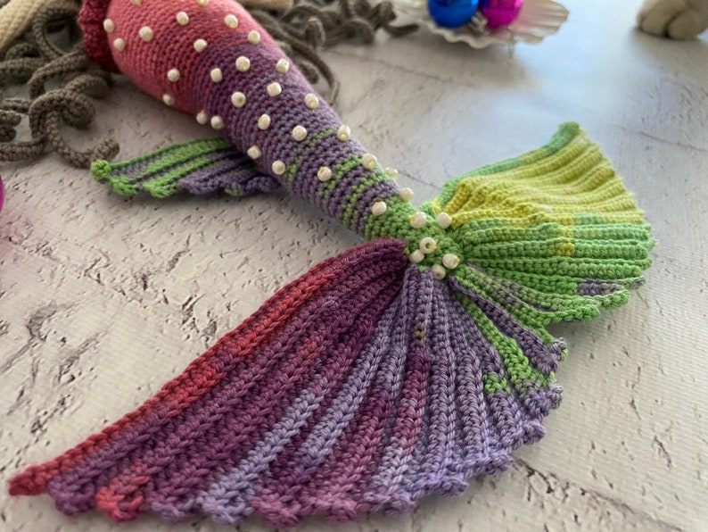May include: A crocheted mermaid tail with a pink, purple, and green color scheme. The tail is decorated with white beads.