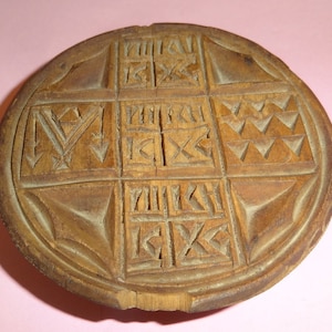 May include: A round wooden stamp with a carved design. The design features a cross in the center with various symbols and letters surrounding it.